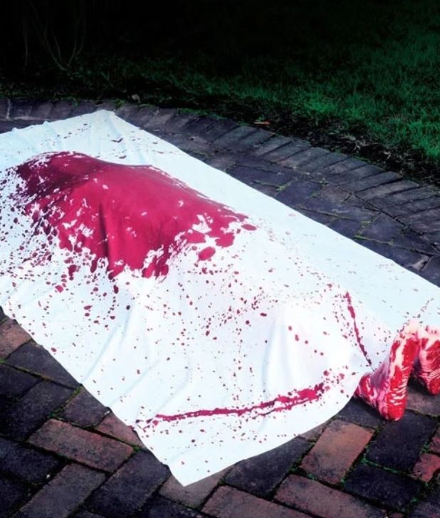 A Halloween yard prop depicting a fake human body lying on a brick patio wrapped in a white sheet stained with heavy red fake blood, with bloody feet extending out from under the sheet. Click to shop all Halloween Blood and Gore.