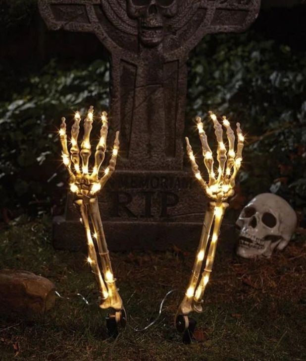 Halloween lawn decoration featuring two illuminated skeleton arm ground stakes glowing with warm lights. They emerge from the ground in front of a stone cross tombstone prop reading "R.I.P." with a skull resting in the background. Click to shop all Halloween Skeleton and Skulls.