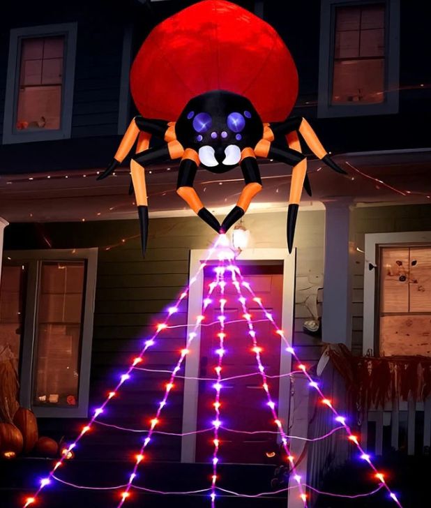 An inflatable black and orange spider with a large red abdomen mounted above a home's front porch, emitting a cone-shaped spiderweb made of purple and red LED string lights stretching down toward the entryway. Click to shop all Halloween Spiders and Web.