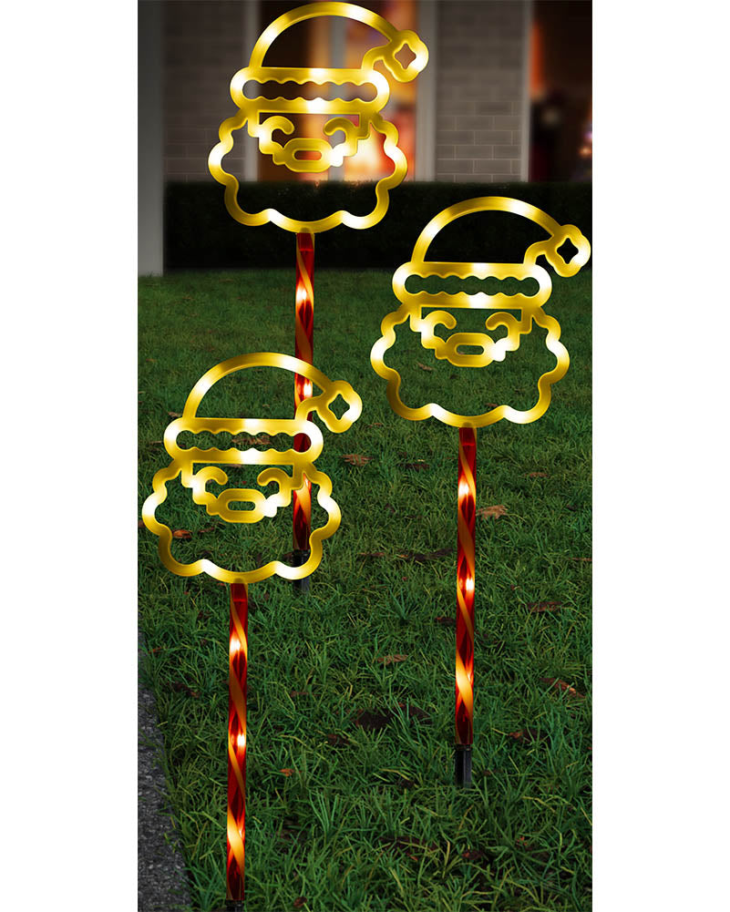 Three gold Santa Claus head yard lights mounted on red-and-gold twisted stake poles, shown lit on a green lawn at night.