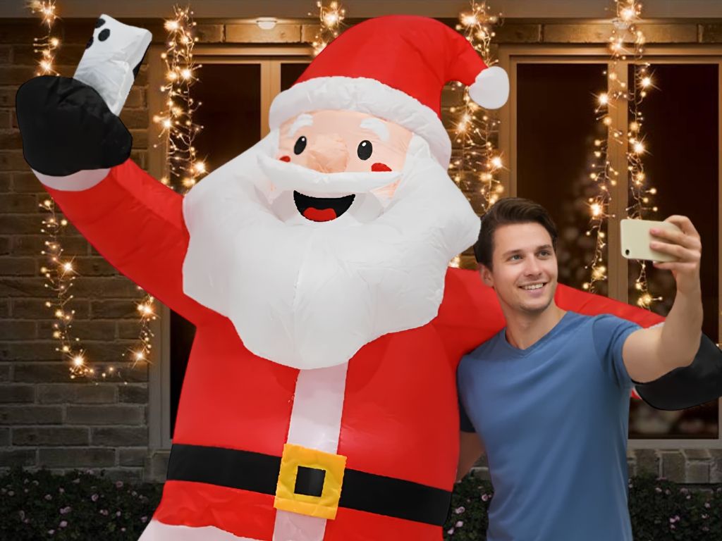 A man taking a selfie with a very large inflatable Santa Claus figure outside a house decorated with warm string lights. Click to shop all Christmas Decorations