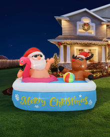 A festive inflatable pool float on a grassy lawn at night in front of a decorated house. The float shows Santa Claus sitting in a pink flamingo-shaped ring, a brown reindeer on the right, and the words 'Merry Christmas' in yellow on a blue pool base, with a small multicolor ball nearby.