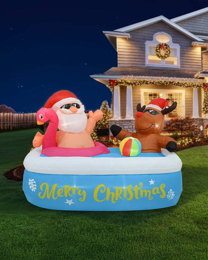 A festive inflatable pool float on a grassy lawn at night in front of a decorated house. The float shows Santa Claus sitting in a pink flamingo-shaped ring, a brown reindeer on the right, and the words 'Merry Christmas' in yellow on a blue pool base, with a small multicolor ball nearby.