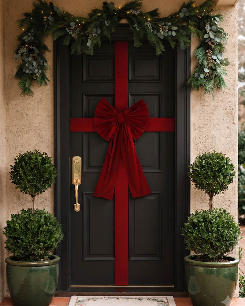 A black front door decorated with a large red ribbon bow and vertical red ribbon, framed by green garland with lights, flanked by two potted green topiary trees.