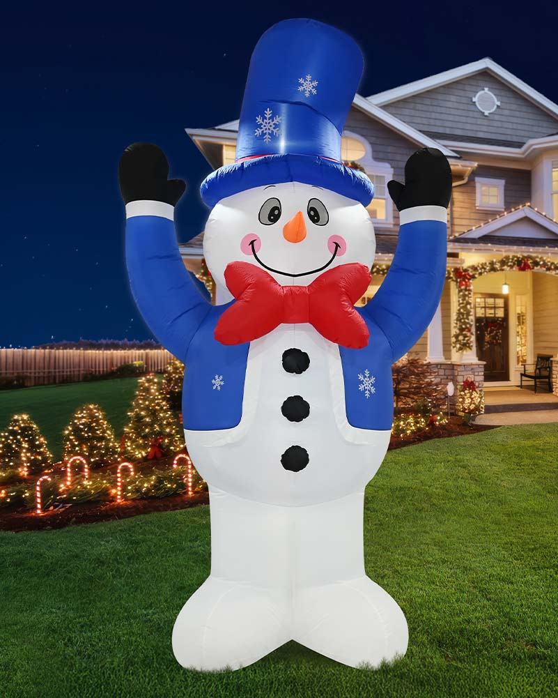 A large inflatable snowman holiday decoration stands on a lawn in front of a decorated house at night. The snowman has a white body, blue jacket and tall blue hat with white snowflake details, a red bow tie, black gloves, and an orange carrot nose.