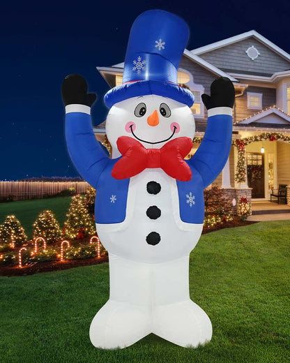A large inflatable snowman holiday decoration stands on a lawn in front of a decorated house at night. The snowman has a white body, blue jacket and tall blue hat with white snowflake details, a red bow tie, black gloves, and an orange carrot nose.