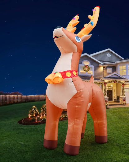 An inflatable Christmas reindeer stands on a green lawn in front of a decorated house at night. The reindeer is tan and cream with orange antlers and a red collar with gold ornaments.