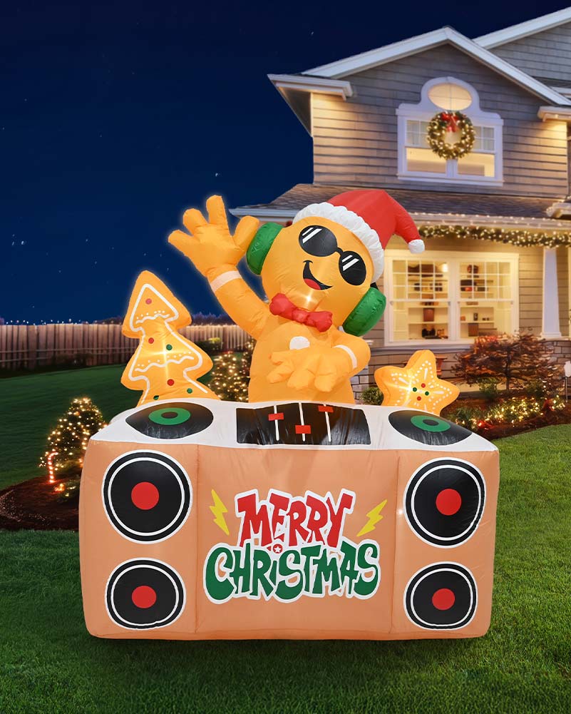 An inflatable Christmas DJ booth decoration stands on a lawn in front of a decorated house at night. The orange character wears a red Santa hat, green headphones, and sunglasses, with a tree and star beside it.