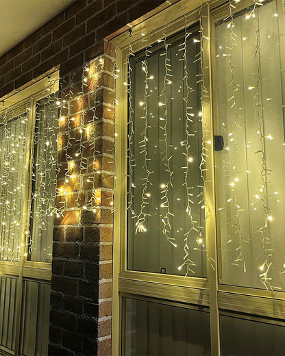 Warm white icicle string lights draped over glass windows and a brick column, creating a cozy festive atmosphere.