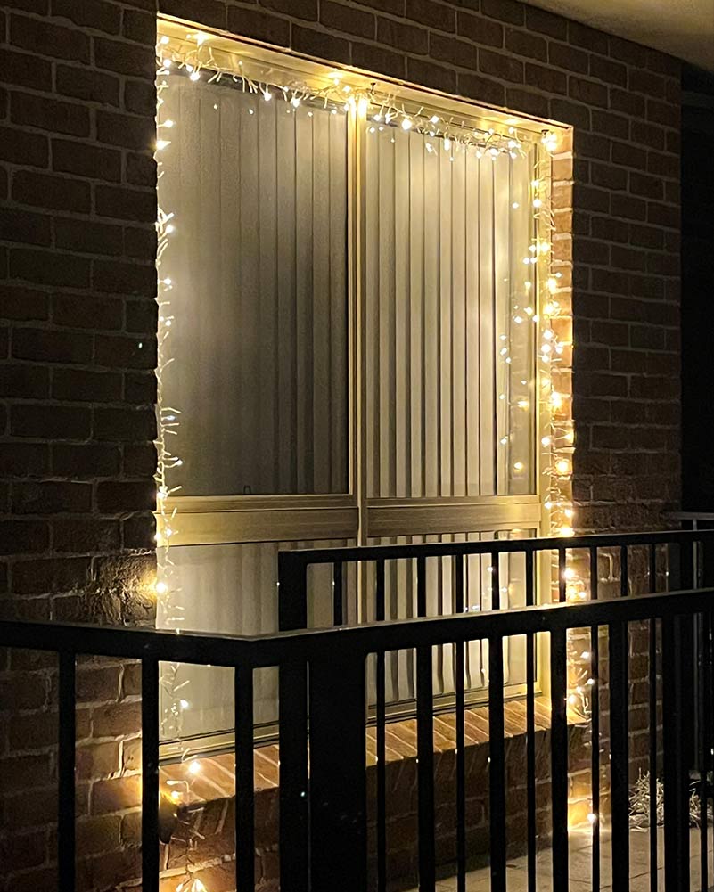 Warm white string lights draped around a sliding glass door on a brick exterior wall, with a black metal railing in the foreground.