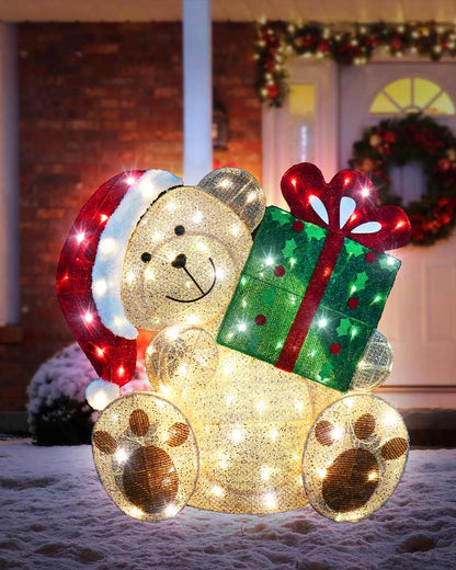 A festive teddy bear holiday light decoration with a Santa hat and a glowing green gift box, displayed outdoors in front of a decorated holiday door.