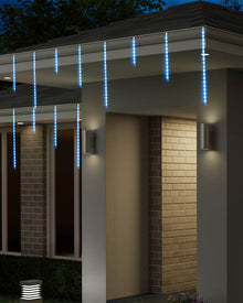 Blue LED icicle string lights hanging from the eaves of a modern house at night, illuminating the entryway and brick columns.