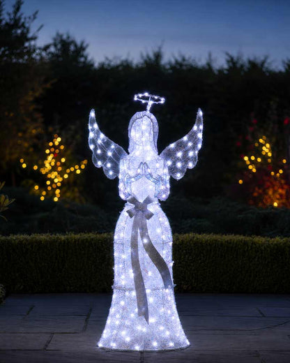 A large illuminated angel garden decoration stands on a patio at dusk, with glowing white lights forming wings, a halo, and a long robe, set against dark hedges and blurred warm string lights.