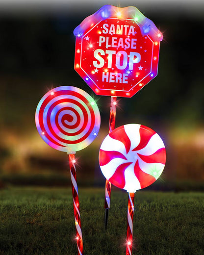 Three festive outdoor yard signs on grass at night: a red stop sign with 'Santa Please Stop Here' and multicolor LED lights, a red-and-white spiral lollipop sign, and a red-and-white swirl candy sign with lights.