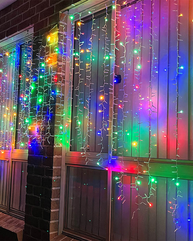 Multicolor Christmas icicle string lights hanging vertically on a screened patio door, casting vibrant rainbow reflections on the glass and wood siding.