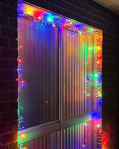 Multicolor LED window and door string lights wrapped around a double-slat metal door set in a brick wall at night.