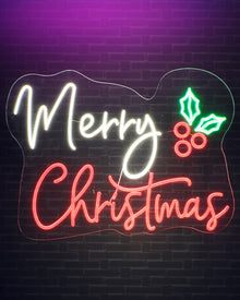 A glowing Christmas neon sign on a dark brick wall, with the words “Merry Christmas” in white and red script and a green holly leaf and berry accent.