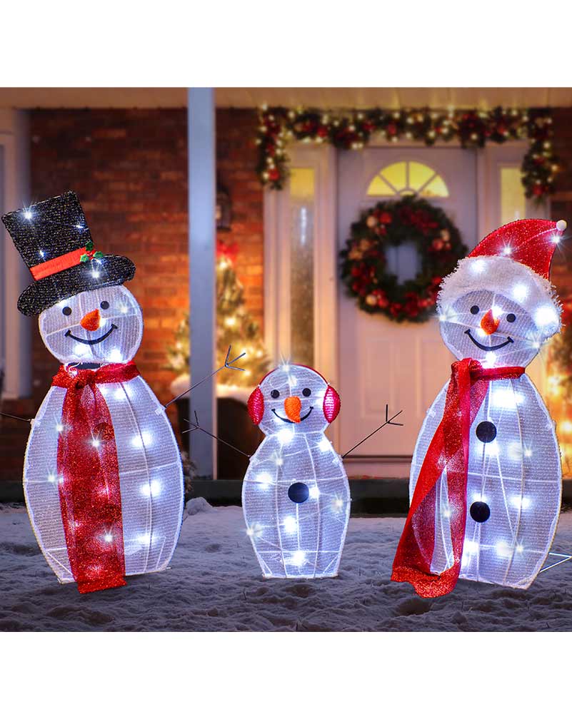 Three illuminated snowman yard decorations in different sizes standing in snow in front of a decorated holiday house entrance. The snowmen are made of white grid material with embedded lights, wearing red scarves, one with a black top hat and one with a red Santa hat.