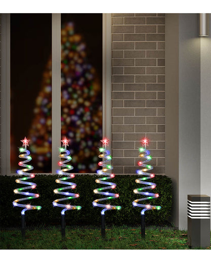 Four spiral Christmas tree light decorations on outdoor stakes, lit with multicolored lights, displayed in front of a house window and brick wall at night.