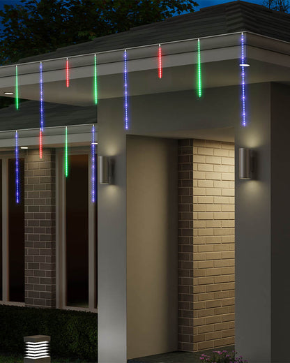 Multicolor LED icicle string lights hanging from the eaves of a modern house at night, casting colorful light beams down the exterior brick walls and entryway.