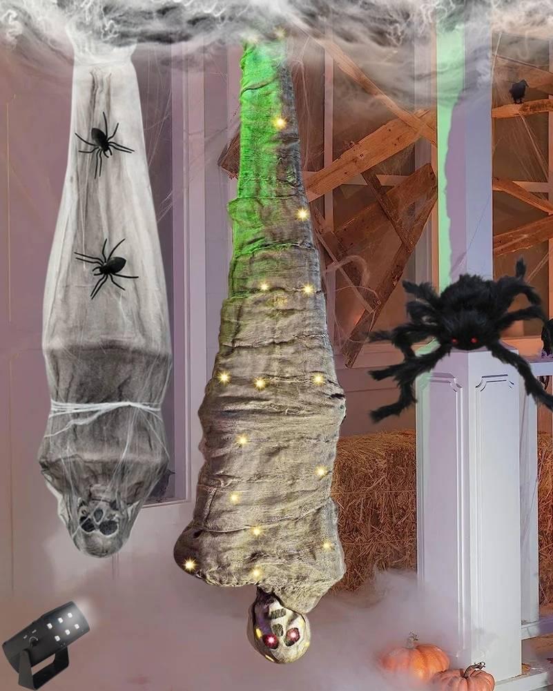 Halloween decoration set featuring a hanging spider web with black spider cutouts, two wrapped corpse props, a large black spider prop, and a flashing light, staged in a decorated indoor room with cobwebs and pumpkins.