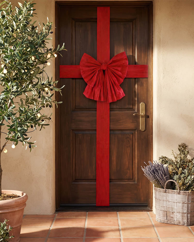 A dark brown wooden front door decorated with a large red fabric bow and matching red ribbon running vertically down the center, shown in a warm beige entryway with potted plants and terracotta tile flooring.