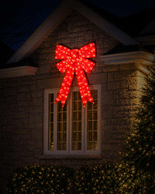 A large red LED light Christmas bow decoration mounted on the gable above a window on a stone house exterior at night.