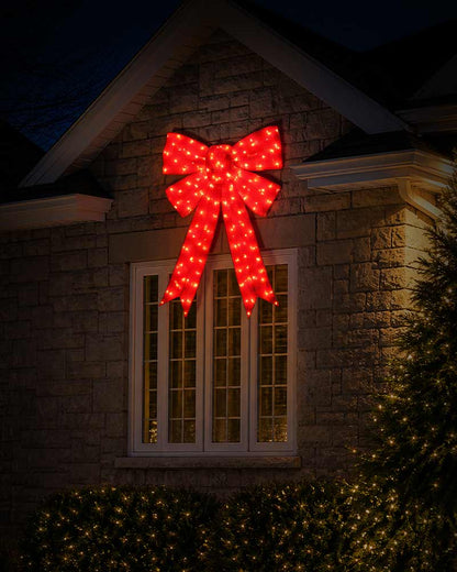 A large red LED light Christmas bow decoration mounted on the gable above a window on a stone house exterior at night.