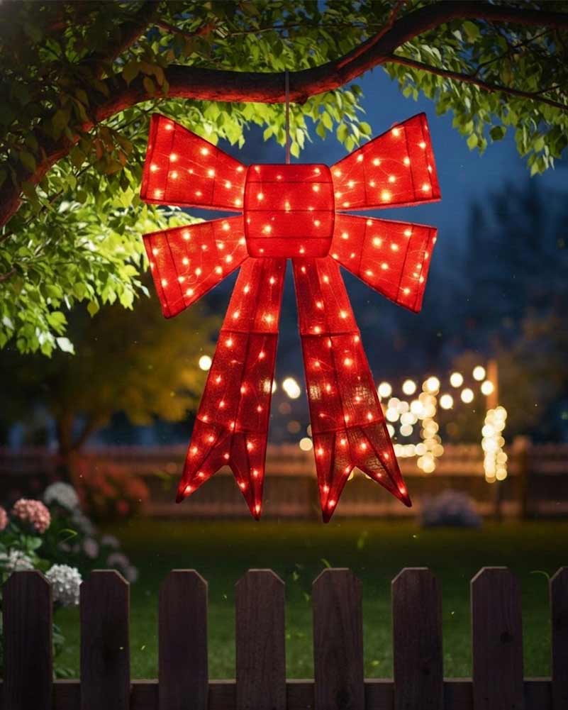 A large red bow-shaped marquee light with glowing warm white LED bulbs hangs from a tree branch outdoors at night, with a blurred garden background and fence in the foreground.