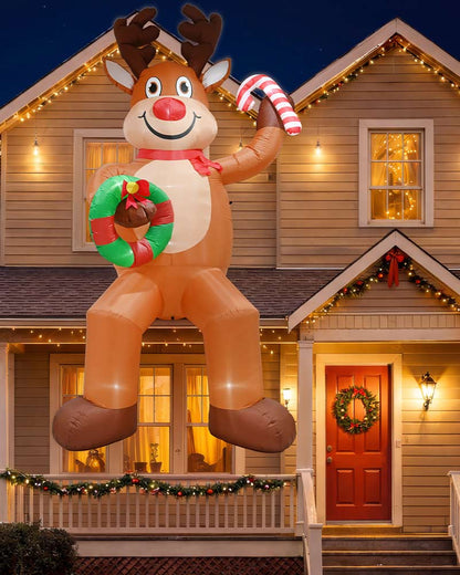 A large inflatable Christmas reindeer decoration stands on a front porch in front of a house at night. The reindeer is tan and white with brown hooves and antlers, holding a green wreath and a red-and-white striped candy cane.