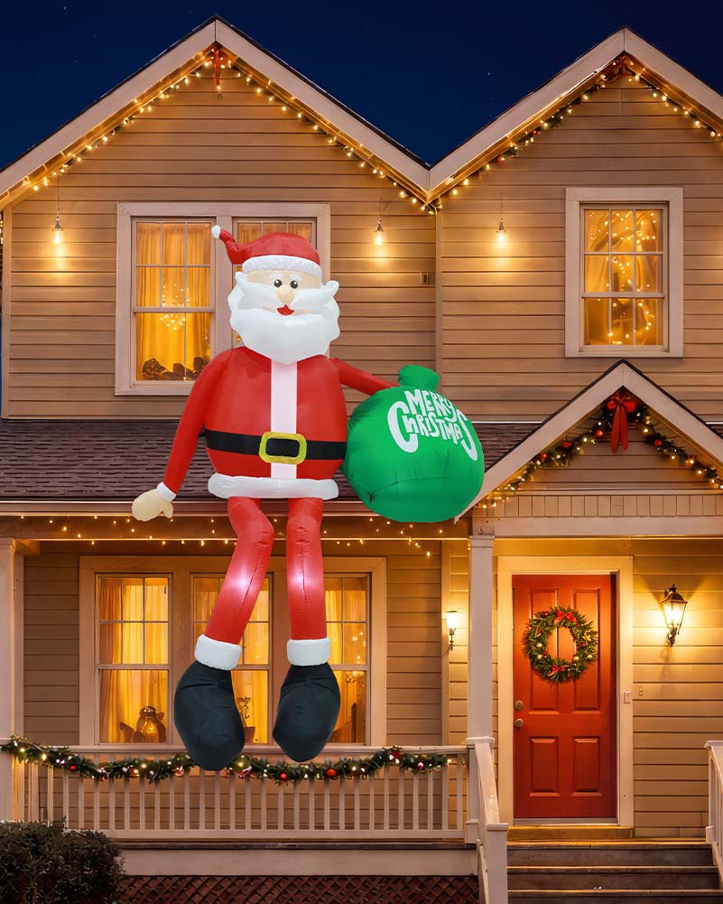 A large inflatable Santa Claus holiday decoration stands on a front porch at night, holding a green sack that reads 'MERRY CHRISTMAS.' The house is lit with warm string lights, and a wreath hangs on the red front door.
