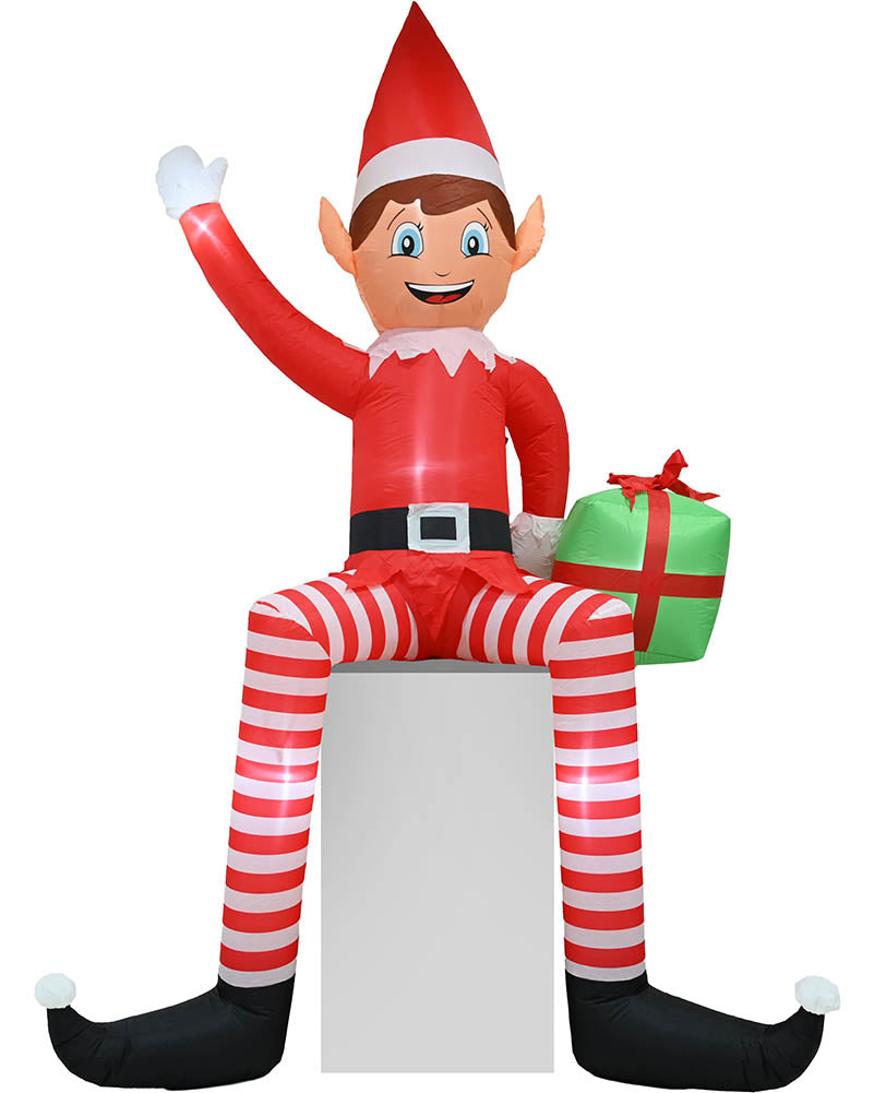 An inflatable Elf on the Shelf Christmas decoration sitting on a white block, wearing a red and white striped outfit with a green gift box, isolated on a white background.
