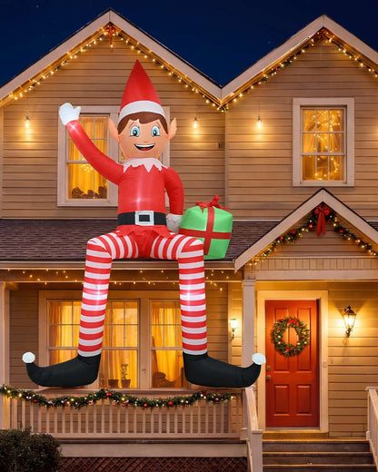 A large inflatable Elf on the Shelf decoration sits on the front porch of a house decorated for Christmas at night. The elf is wearing a red suit with white striped leggings, black shoes, and is holding a green gift box with a red bow.