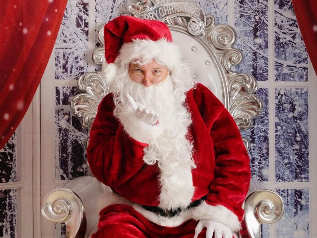 Santa Claus wearing round spectacles and white gloves, sitting on an ornate silver throne against a snowy winter window backdrop. Click to shop all Santa Suits.