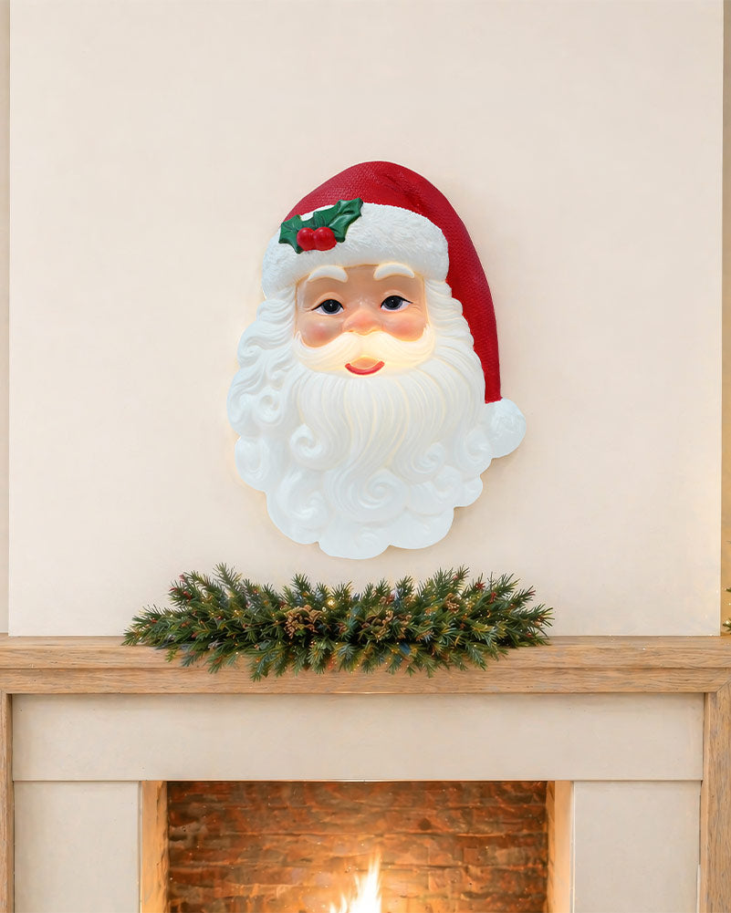A Santa Claus face wall light with a glowing white beard and red hat, mounted above a fireplace mantel with a green garland.