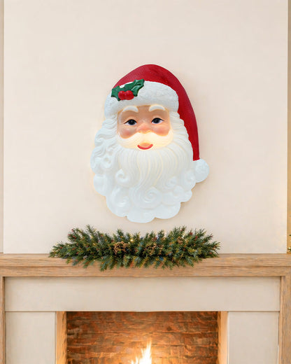 A Santa Claus face wall light with a glowing white beard and red hat, mounted above a fireplace mantel with a green garland.