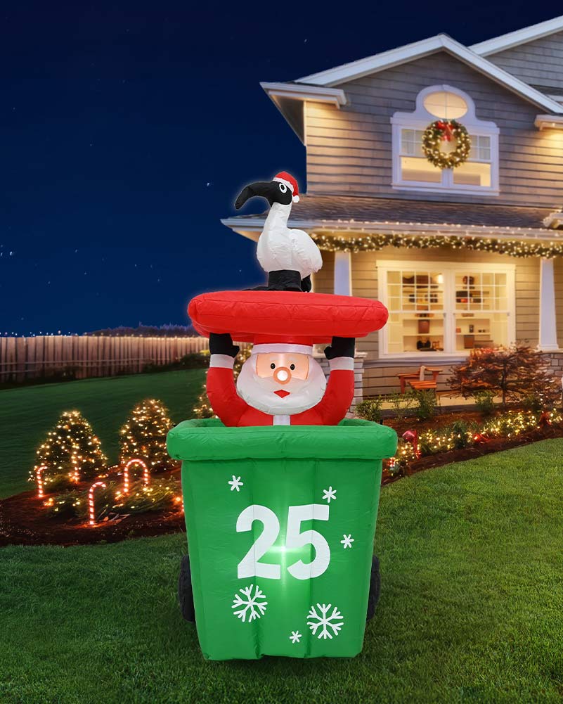 A large inflatable Christmas yard decoration stands on a lawn in front of a decorated house at night. The design shows Santa Claus emerging from a green trash can with the number 25 on the front, topped with a red hat and a white bird perched on his shoulder.