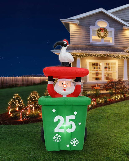 A large inflatable Christmas yard decoration stands on a lawn in front of a decorated house at night. The design shows Santa Claus emerging from a green trash can with the number 25 on the front, topped with a red hat and a white bird perched on his shoulder.