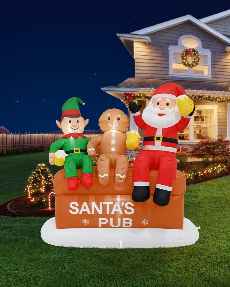 A large inflatable Christmas yard sign shaped like Santa's Pub, featuring Santa Claus, an elf, and a gingerbread man sitting on an orange crate. The sign is displayed on a green lawn in front of a decorated house at night.