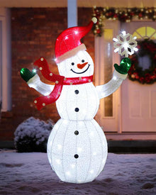 A large snowman holiday light decoration stands in front of a house door. The snowman is white with glowing lights, wearing a red hat, red scarf, green gloves, and holding a white snowflake.