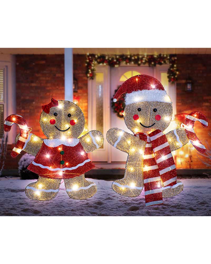 Two glittery Christmas yard signs shaped like a gingerbread woman and a gingerbread man, decorated with warm white lights and red-and-white holiday clothing, standing in snow in front of a decorated house.