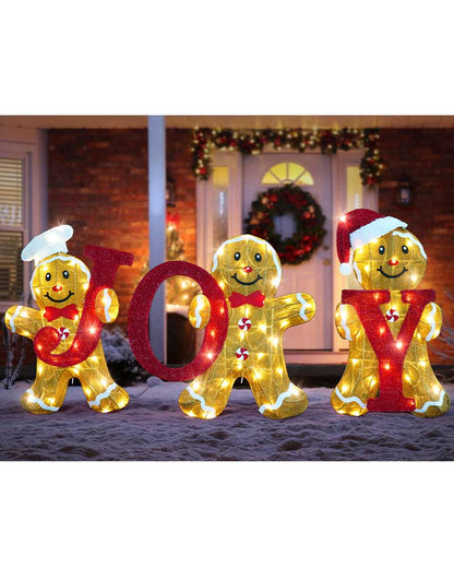 Three gingerbread man holiday light decorations spelling JOY in front of a decorated house door at night.