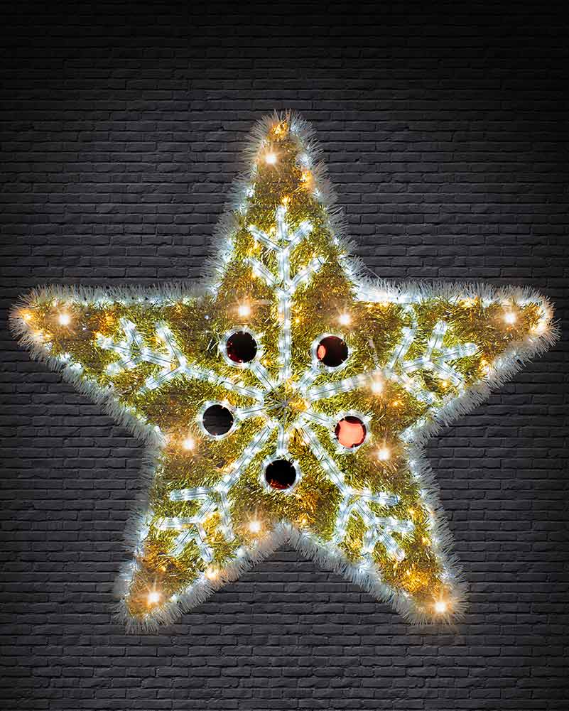 A five-pointed star-shaped Christmas decoration made of green pine branches, illuminated with warm yellow and cool white lights, set against a dark gray brick wall.