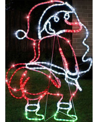 A large outdoor Christmas light decoration shaped like Santa Claus, outlined in red, white, and green LED lights, photographed at night on grass in front of a fence and brick wall.