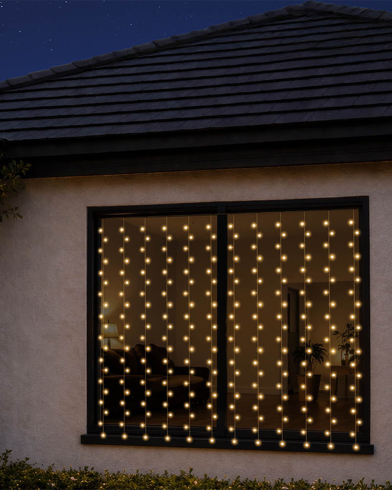 Warm white curtain string lights hanging in vertical strands across a large window on a house exterior at night.