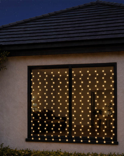 Warm white curtain string lights hanging in vertical strands across a large window on a house exterior at night.