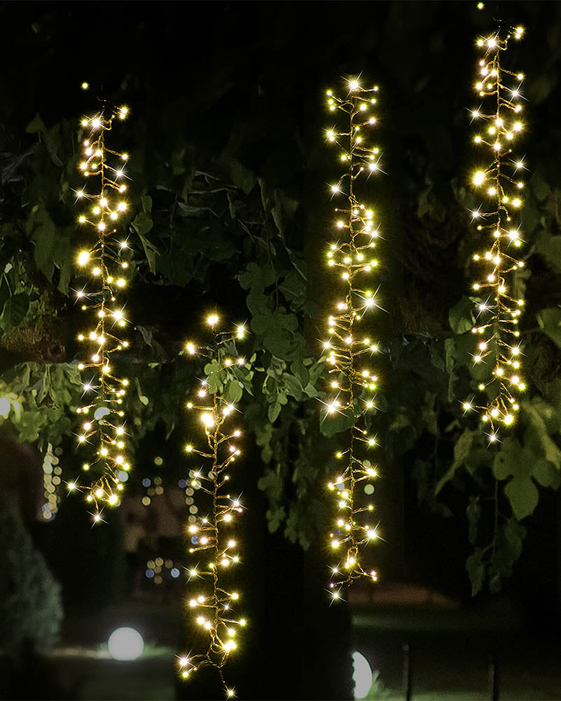 Warm white string lights draped vertically over dark tree branches and leaves at night, creating bright glowing strands against a dark outdoor background.