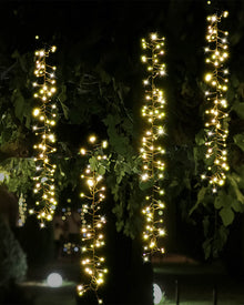 Warm white string lights draped vertically over dark tree branches and leaves at night, creating bright glowing strands against a dark outdoor background.