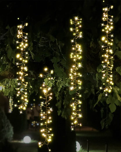 Warm white string lights draped vertically over dark tree branches and leaves at night, creating bright glowing strands against a dark outdoor background.