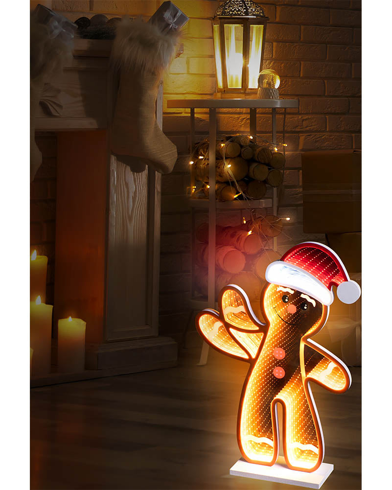 A glowing Christmas gingerbread man light figure wearing a red Santa hat, placed on a white base in a warmly lit holiday room with candles, a lantern, and stacked logs in the background.